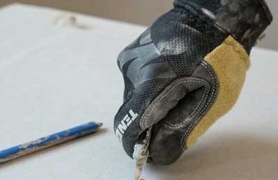 A plasterer wearing a glove cutting plasterboard with a knife and ruler